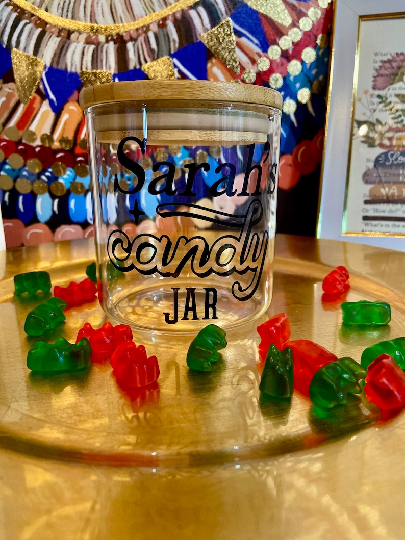 May include: A glass candy jar with a bamboo lid. The jar has a black vinyl decal that says "Sarah's candy JAR". The jar is surrounded by red and green gummy bears.