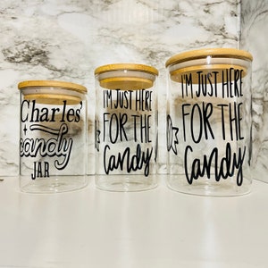 May include: Three clear glass jars with bamboo lids. The jars have black text that reads "Charles candy jar" and "I'M JUST HERE FOR THE Candy". The jars are different sizes and are arranged in a row.