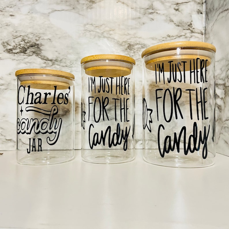 May include: Three clear glass jars with bamboo lids. The jars have black text that reads "Charles candy jar" and "I'M JUST HERE FOR THE Candy". The jars are different sizes and are arranged in a row.