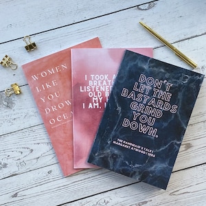 Peut inclure: Trois carnets avec des citations inspirantes. L'un est corail avec le texte "WOMEN LIKE YOU DROW OCEAN". Un autre est rose avec le texte "I TOOK A BREATH LISTENED OLD B MY I AM, I". Le troisième est bleu foncé avec le texte "DON'T LET THE BASTARDS GRIND YOU DOWN."