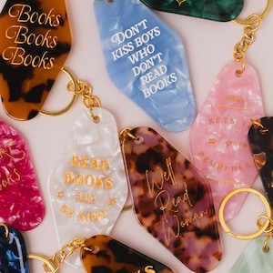 Pode incluir: Uma coleção de porta-chaves estilo motel em acrílico marmoreado de várias cores, incluindo tartaruga, rosa, azul e branco. Cada porta-chaves possui argolas e correntes de metal dourado, com textos gravados como Books, Read Books e Reading is sexy.