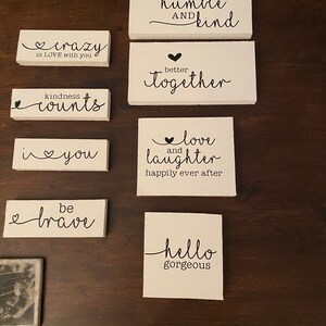 May include: A set of ten white wooden signs with black lettering. The signs feature various inspirational quotes and sayings, such as "always stay humble and kind", "better together", "love and laughter", and "hello gorgeous".