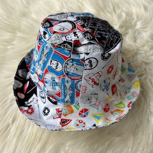 May include: A reversible bucket hat featuring a patchwork design with Star Wars-themed graphics. The hat showcases iconic characters and symbols in red, blue, black, and white. The brim and crown are divided into sections with different patterns.