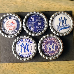 May include: Five round, silver-colored bottle caps with different New York Yankees logos. The logos feature the team's iconic 'NY' logo in various styles, including a classic baseball cap, a vintage-inspired design, and a modern, bold font. The caps are perfect for adding a touch of team spirit to any space.