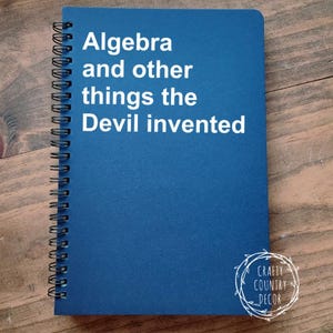 May include: A blue spiral-bound notebook with the text "Algebra and other things the Devil invented" in white. The notebook has a Crafty Country Decor logo in the bottom right corner. The notebook is on a wooden surface.