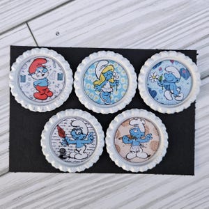 May include: Five white bottle caps with Smurf characters. Each cap features a different Smurf illustration, including Smurfette and other Smurfs. The caps are arranged on a black surface, showcasing the colorful designs.