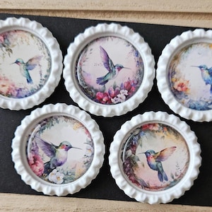 May include: Five white bottle caps with a floral design and a hummingbird painted on each. The hummingbirds are painted in shades of blue, purple, and green.