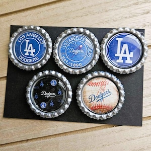 May include: Five bottle caps with the Los Angeles Dodgers logo and baseball imagery. The caps feature the team's blue and white colors, with the "LA" logo and the word "Dodgers" prominently displayed. The caps are arranged on a black surface.