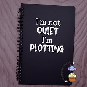 May include: Black spiral-bound notebook with the text "I'm not QUIET I'm PLOTTING" in white. A small illustration of a cartoon bee with flowers is in the bottom right corner. The notebook is on a light brown wooden surface.