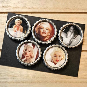 May include: Five silver bottle cap magnets with different images of Marilyn Monroe. Each magnet features a different photo of the iconic actress.