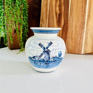 May include: A white ceramic vase with a blue and white windmill scene. The vase has a round body and a narrow neck. The scene depicts a windmill, houses, and a river.