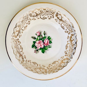 May include: A white ceramic plate with a gold rim and decorative gold filigree around the edge. The centre has a bouquet of pink roses and green leaves. A classic vintage design.