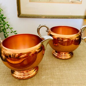 May include: A set of two copper sugar and creamer bowls with silver handles. The bowls are round and have a shiny finish.