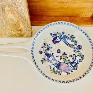 May include: A white ceramic serving dish with a blue and green floral design featuring two figures. The dish has a handle and a scalloped edge.