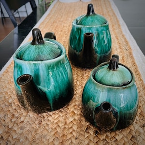 Puede incluir: Tres teteras de cerámica con esmalte verde y negro. Las teteras tienen tapas y están sobre una estera tejida.