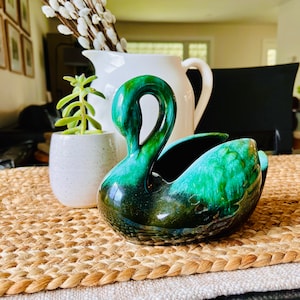 Puede incluir: Una figurita de cisne de cerámica verde y negra esmaltada. El cisne tiene un cuello largo y una envergadura amplia. La figurita está sentada sobre un mantel individual de paja trenzada.