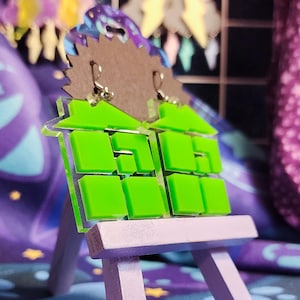 以下が含まれることがあります： ネオンの緑とグレーの幾何学的なイヤリングのペア。イヤリングは、正方形の窓が付いた様式化された家の形をしています。カラフルな背景に対して、小さな薄紫色のイーゼルに表示されています。