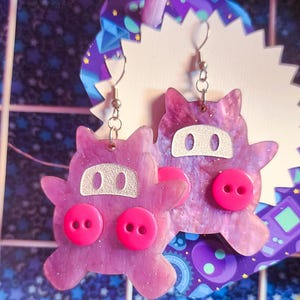 May include: A pair of handmade earrings featuring a whimsical purple creature design. The earrings have a glittery finish, silver-tone ear wires, and bright pink button accents for eyes and a nose. The earrings are displayed against a patterned background.