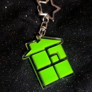 以下が含まれることがあります： 銀色の星型の留め金が付いた、ネオングリーンの幾何学的な家の形をしたキーホルダー。キーホルダーは透明な縁取りと、一連の緑色の正方形と長方形があります。背景は、金色のグリッターが付いた黒い生地です。