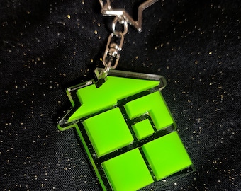 May include: A neon green geometric house-shaped keyring with a silver star-shaped clasp. The keyring has a clear border and a series of green squares and rectangles. The background is a black fabric with gold glitter.