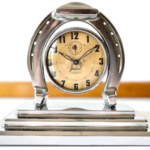 May include: A silver horseshoe-shaped clock with a white face and black hands. The clock is on a silver stand with a stepped design. The clock face reads "Juliet The Clock".
