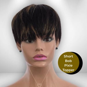 May include: A mannequin head displaying a short, layered bob pixie topper wig. The wig is black with subtle blonde highlights. The mannequin wears a gold heart pendant necklace and black stud earrings. The text "Short Bob Pixie Topper" is in a gold circle.