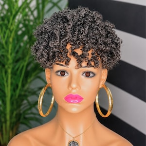 May include: A mannequin head featuring a short, dark, tightly coiled curly wig with a textured finish. The mannequin has brown eyes, bright pink lips, large gold hoop earrings, and a gold chain necklace with a circular pendant.