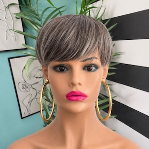 May include: A mannequin head with a short, layered, salt-and-pepper gray pixie haircut. The mannequin features dark brown eyes, bright pink lips, and large, smooth, circular gold-tone hoop earrings that hang from the earlobes.