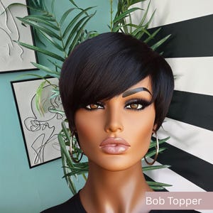 May include: A black bob wig on a mannequin with hoop earrings. The mannequin has brown eyes, full lips, and dramatic eye makeup. The background features a black and white striped pattern and a teal wall with artwork and greenery. The text "Bob Topper" is visible.