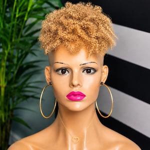 May include: A mannequin head featuring short, curly, light brown hair. The mannequin has bright pink lips, large gold hoop earrings, and a thin gold necklace with a small heart-shaped pendant. The skin tone is light tan with defined facial features.