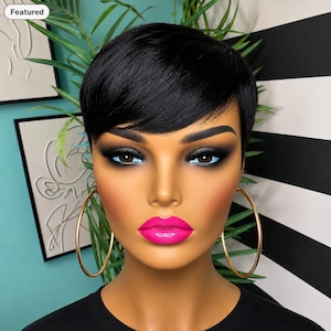 May include: A mannequin head with short, straight black hair, dark eye makeup, and bright pink lipstick. The mannequin wears large, thin gold hoop earrings and a plain black shirt. The facial features are smooth with a tan complexion.