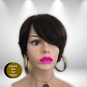 May include: A layered bob hair topper in dark brown, styled with a side-swept bang. The mannequin head features large gold hoop earrings and a necklace with a pendant. The lips are painted a bright pink. The text "Layered Bob Hair Topper" is in a circle.