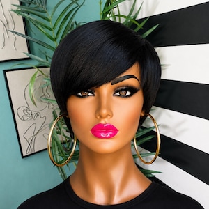 May include: A mannequin head featuring a short, straight black wig with side-swept bangs. The mannequin has brown eyes, long dark eyelashes, and bright pink glossy lips. Large gold-toned hoop earrings are attached to the mannequin's ears.