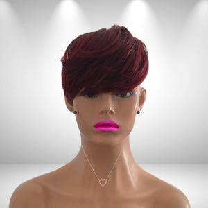 May include: A mannequin head with a short, layered burgundy wig. The mannequin has dark eyes, black stud earrings, and bright pink lipstick. A delicate gold necklace with a heart pendant is visible.