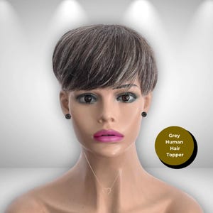 May include: A mannequin head displaying a short, layered grey human hair topper. The hair is styled with a side-swept bang. The mannequin has dark eyes, pink lipstick, and black stud earrings. A gold-colored heart pendant necklace is visible. A circular label reads "Grey Human Hair Topper."