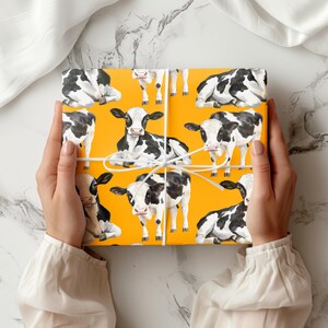 May include: Gift wrap featuring a repeating pattern of black and white cows on a bright yellow background. The cows are in various poses. The gift wrap is tied with white string.