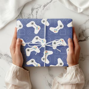 May include: A wrapped present with a blue background featuring a pattern of white video game controllers and other gaming icons. The gift is tied with white string and is held by a person. The wrapping paper has a repeating pattern of controllers.