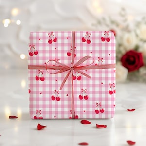 May include: A gift box wrapped in pink and white gingham paper, decorated with red cherries and hearts. A red ribbon is tied in a bow around the box. The box sits on a white surface, scattered with red rose petals.