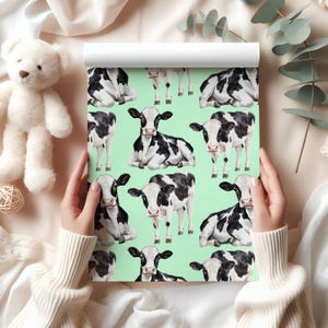May include: A roll of wallpaper featuring a repeating pattern of black and white cows on a light green background. The cows are in various poses, some standing and others lying down. The wallpaper is held by a person wearing a white sweater.