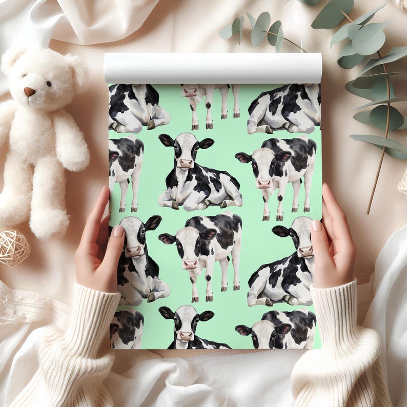 May include: A roll of wallpaper featuring a repeating pattern of black and white cows on a light green background. The cows are in various poses, some standing and others lying down. The wallpaper is held by a person wearing a white sweater.