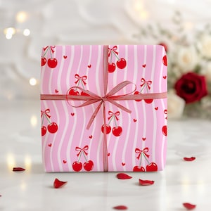 Könnte beinhalten: Ein Geschenkbox, verpackt in rosa Papier mit einem Muster aus roten Kirschen und weißen, wellenförmigen Streifen. Die Box ist mit einem weinroten Band und einer Schleife gebunden. Kleine rote Herzen sind auf dem Papier verstreut.