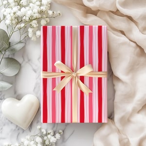Könnte beinhalten: Eine rechteckige Geschenkbox, verpackt in rot-, rosa- und weiß gestreiftem Papier, gebunden mit einem goldenen Satinband und einer Schleife. Die Box befindet sich auf einer Marmoroberfläche, mit weißen Blumen und einem herzförmigen Objekt.