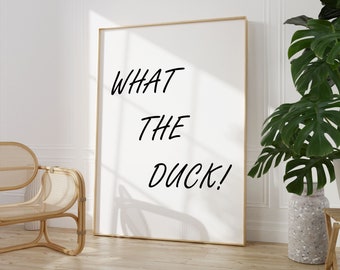 May include: A white framed poster with the text "WHAT THE DUCK!" in black cursive font. The poster is hanging on a white wall in a room with a wicker chair and a potted plant.