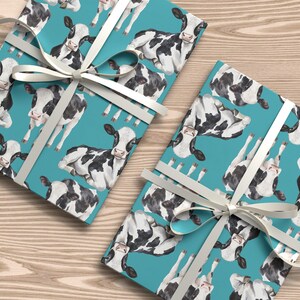 May include: Two wrapped gifts with a teal background featuring a pattern of black and white cows. The gifts are tied with white ribbon and bows. The gifts are on a light brown wooden surface.