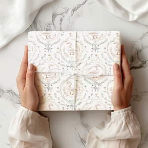 May include: A square gift box wrapped in white paper featuring a repeating pattern of light brown geese surrounded by green botanical wreaths and small blue flowers, tied with a thin white string in a centred bow across the front surface.