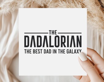 Könnte beinhalten: Eine weiße quadratische Karte mit dem Text "THE DADALORIAN THE BEST DAD IN THE GALAXY" in Schwarz. Die Karte wird von einer Person mit neutralem Hintergrund gehalten.