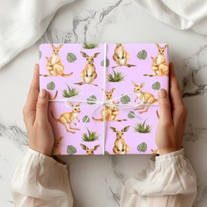 May include: A wrapped gift with a repeating pattern of kangaroos, baby kangaroos, and tropical foliage on a pale purple background. The gift is tied with white twine. The theme is nature-inspired gift wrap.
