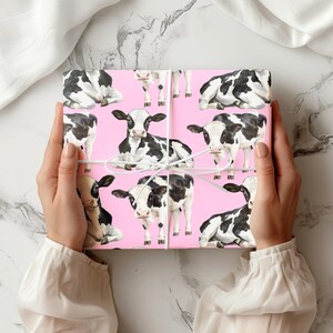 May include: A pink gift box wrapped with paper featuring a pattern of black and white cows. The box is tied with white string. The background is a marble pattern.