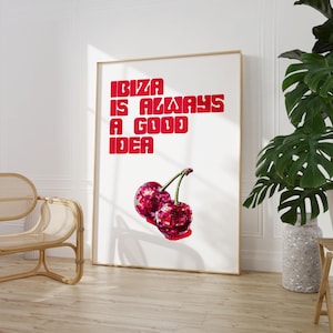 May include: A framed art print with the text "IBIZA IS ALWAYS A GOOD IDEA" in red block letters. Below the text are two cherries with disco ball textures. The print is in a room with a wooden floor and a potted plant.