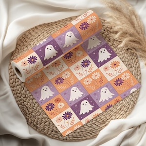 Könnte beinhalten: Eine Rolle gemustertes Papier mit einem sich wiederholenden Design aus lächelnden weißen Geistern und Blumenarrangements in einem Rastermuster. Die Quadrate wechseln sich zwischen orange, lila und cremefarben ab. Das Papier wird auf einem Weidenkorb präsentiert.